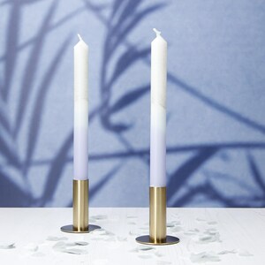 May include: Two white candles with a gradient of purple to white, in gold candle holders on a white surface.