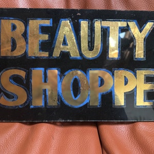 Puede incluir: Un letrero de vidrio negro con letras doradas que dice "BEAUTY SHOPPE". Las letras están delineadas en azul.