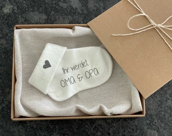 Op de afbeelding: Een witte babysok met een grijs hart en de tekst "Ihr werdet OMA & OPA" in een bruine cadeaudoos met een beige stoffen voering.