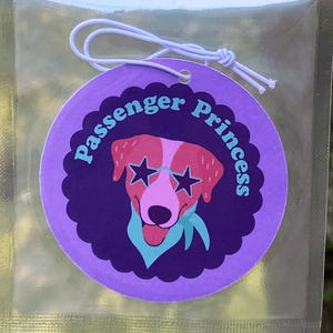 May include: Air freshener car charm with ocean scent. The packaging features a purple circular charm with a dog wearing star-shaped sunglasses and the words "Passenger Princess". The charm is in a clear plastic bag.