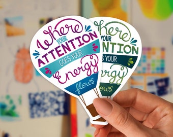 May include: Two hot air balloon-shaped stickers with the text "Where your attention goes your energy flows." One sticker has purple and teal accents, the other has green and teal accents. The stickers are held by a hand.