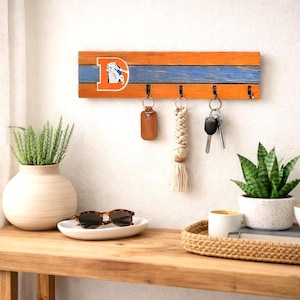 May include: A wooden key holder with an orange and blue color scheme, featuring the Denver Broncos logo. It has four hooks with keys and keychains hanging from them. The key holder is mounted on a white wall.