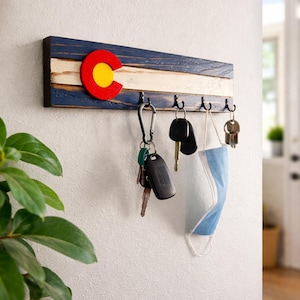 May include: A wooden key holder featuring the Colorado state flag design. The rectangular board has a blue, white, and dark wood grain pattern with a red and yellow emblem. Several keys and a face mask hang from the hooks.