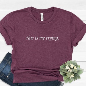 May include: A maroon t-shirt with the text "this is me trying." printed in white on the front.
