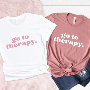 May include: Two t-shirts with the text "go to therapy." printed in pink on a white shirt and white on a pink shirt.
