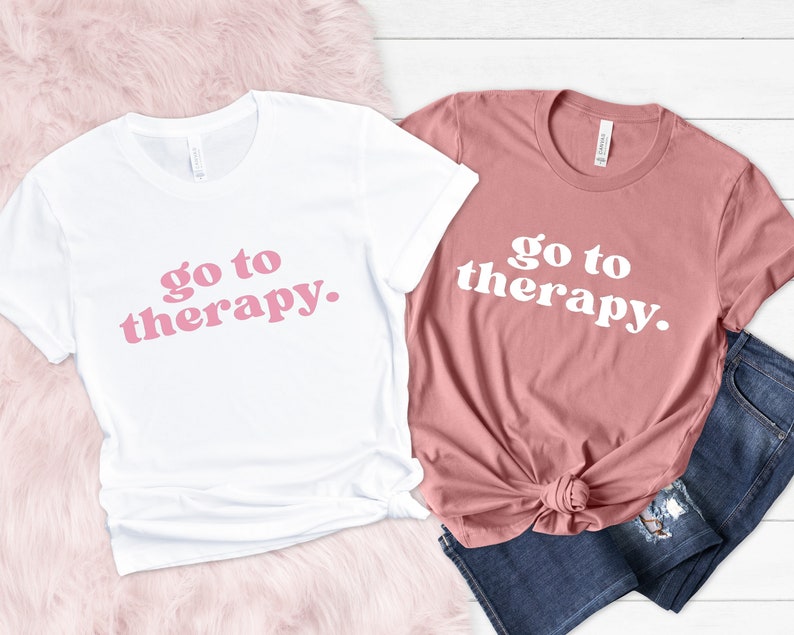 May include: Two t-shirts with the text "go to therapy." printed in pink on a white shirt and white on a pink shirt.