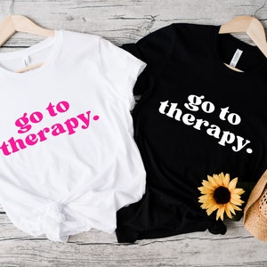 May include: Two t-shirts with the text "go to therapy." One shirt is white with pink text and the other is black with white text.