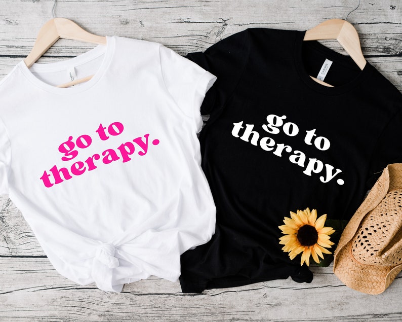 May include: Two t-shirts with the text "go to therapy." One shirt is white with pink text and the other is black with white text.