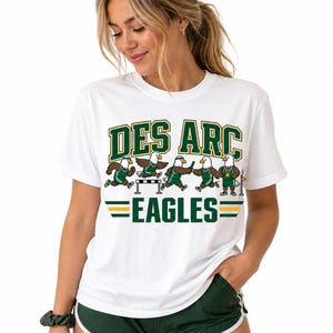 May include: A white short-sleeve t-shirt featuring the text DES ARC in green with gold outlines above a graphic of five cartoon eagles in green and gold track uniforms participating in various track and field events, with EAGLES below.