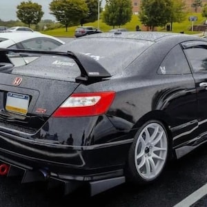 May include: A black Honda Civic coupe with a large rear wing, white wheels, and a red tow hook. The car is parked in a car park and has rain drops on the body.
