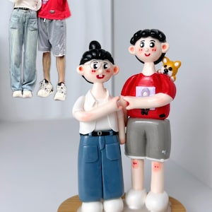 Può includere: Una statuina fatta a mano di una coppia in piedi insieme. La donna indossa una camicia bianca, jeans blu e occhiali. L'uomo indossa una camicia rossa, pantaloncini grigi e ha un piccolo cane sulla spalla. Le statuine sono in piedi su una base di legno.