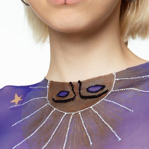 May include: Purple mesh top with a brown and black embroidered sun design with white rays. The sun has two blue eyes.
