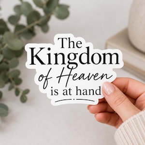 May include: A white die-cut sticker with a black serif and script font that reads The Kingdom of Heaven is at hand. The sticker features a decorative horizontal line with a small dot at the bottom center, creating a clean and legible design.