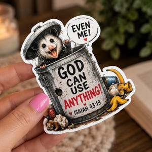 May include: A die-cut sticker featuring a grinning opossum peeking out of a metal trash can. The can displays the text GOD CAN USE ANYTHING! and ISAIAH 43:19, with a speech bubble above the opossum saying EVEN ME! and a small red heart.