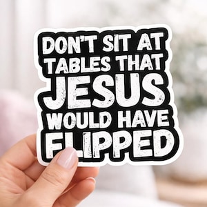 May include: A white sticker with a black border and text that reads "DON'T SIT AT TABLES THAT JESUS WOULD HAVE FLIPPED." The sticker is held by a hand.