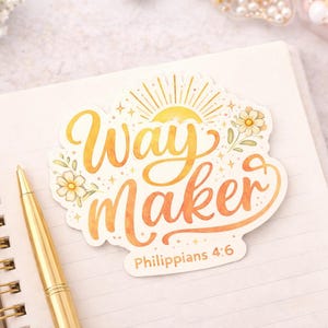 May include: A sticker with the words "Way Maker" in orange and yellow script, with a sun and floral design. The text "Philippians 4:6" is below the main text. The sticker is on a white notebook with a gold pen.