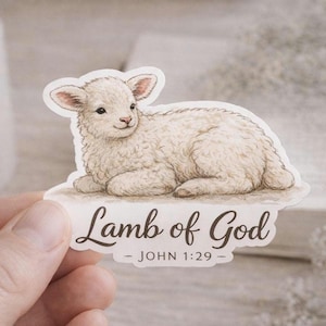 May include: A sticker featuring a watercolor illustration of a fluffy white lamb. The text "Lamb of God - JOHN 1:29" is written in elegant script below the lamb. The sticker has a white border and is held by a hand.