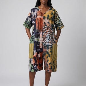 May include: A knee-length jumpsuit featuring a V-neckline and short sleeves, decorated with a multi-colored abstract tie-dye pattern in shades of orange, yellow, green, and black. The garment includes two large, vertical-striped olive green pockets.