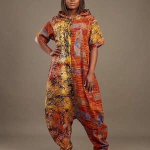 May include: A hooded jumpsuit with a vibrant, abstract pattern in shades of orange, yellow, and black. The short-sleeved garment features a relaxed fit and is paired with white sneakers. The design incorporates a mix of geometric and organic shapes.
