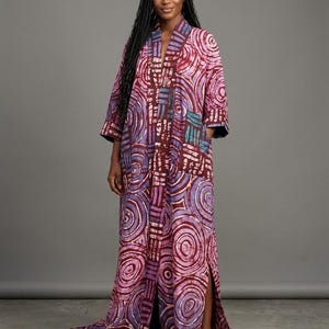 May include: A floor-length, open-front robe in shades of pink, purple, and burgundy, featuring a repeating circular pattern. The robe has three-quarter sleeves and side slits, creating a flowing silhouette. The fabric appears to be a soft, lightweight material.