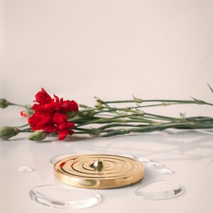 K&ouml;nnte beinhalten: Eine goldene Metallscheibe mit einer erhabenen Mitte und konzentrischen Kreisen. Die Scheibe liegt auf einer wei&szlig;en Oberfl&auml;che mit klaren Wassertropfen darum herum. Ein Strau&szlig; roter Nelken liegt links von der Scheibe.