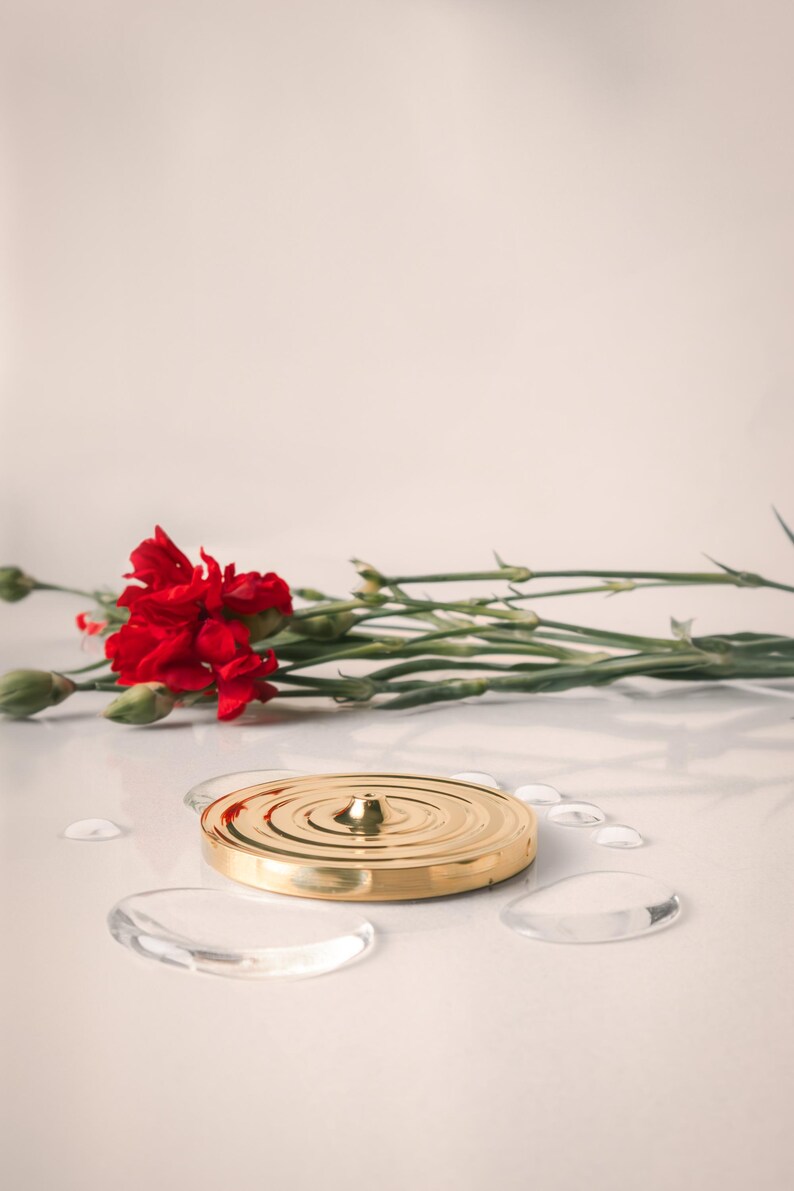 K&ouml;nnte beinhalten: Eine goldene Metallscheibe mit einer erhabenen Mitte und konzentrischen Kreisen. Die Scheibe liegt auf einer wei&szlig;en Oberfl&auml;che mit klaren Wassertropfen darum herum. Ein Strau&szlig; roter Nelken liegt links von der Scheibe.