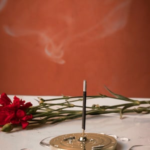 K&ouml;nnte beinhalten: Ein goldfarbener Metall-R&auml;ucherofen mit einem einzigen brennenden R&auml;ucherst&auml;bchen, das Rauch abgibt. Der R&auml;ucherofen steht auf einer wei&szlig;en Oberfl&auml;che mit einer roten Blume und gr&uuml;nen Stielen im Hintergrund.