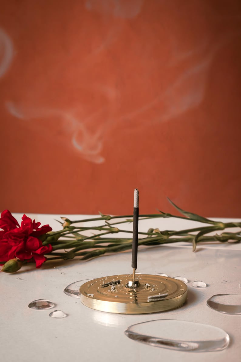 K&ouml;nnte beinhalten: Ein goldfarbener Metall-R&auml;ucherofen mit einem einzigen brennenden R&auml;ucherst&auml;bchen, das Rauch abgibt. Der R&auml;ucherofen steht auf einer wei&szlig;en Oberfl&auml;che mit einer roten Blume und gr&uuml;nen Stielen im Hintergrund.