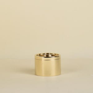 May include: A gold-coloured, cylindrical container with a tiered, circular design on top. The container has a smooth, reflective surface and a small, recessed area at the top. The overall design is minimalist and modern.