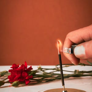 K&ouml;nnte beinhalten: Eine Hand z&uuml;ndet einen schwarzen R&auml;ucherst&auml;bchen mit einem silbernen Feuerzeug an. Der R&auml;ucherst&auml;bchen befindet sich auf einem goldenen Metall-R&auml;ucherst&auml;bchenhalter. Im Hintergrund sind eine rote Nelke und gr&uuml;ne Bl&auml;tter zu sehen.