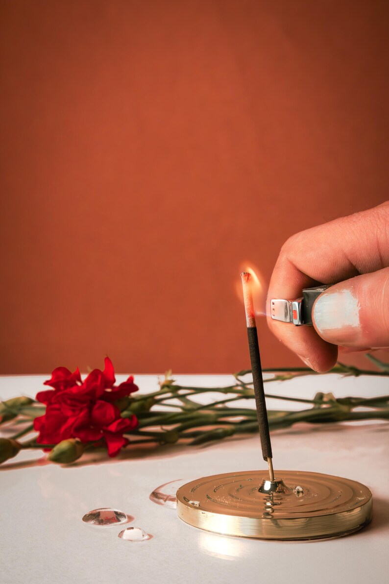 K&ouml;nnte beinhalten: Eine Hand z&uuml;ndet einen schwarzen R&auml;ucherst&auml;bchen mit einem silbernen Feuerzeug an. Der R&auml;ucherst&auml;bchen befindet sich auf einem goldenen Metall-R&auml;ucherst&auml;bchenhalter. Im Hintergrund sind eine rote Nelke und gr&uuml;ne Bl&auml;tter zu sehen.