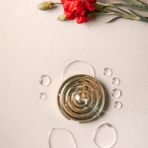 K&ouml;nnte beinhalten: Ein goldfarbener Metall-R&auml;ucherofen mit einem runden Design und einem kleinen Loch in der Mitte. Der R&auml;ucherofen ist von Wassertropfen auf einer hellbeigen Oberfl&auml;che umgeben.