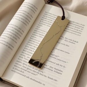 May include: A gold-toned metal bookmark with a reflective surface and a thin, engraved line map design. The bookmark features a dark brown cord attached to the top and is placed between the pages of an open book with printed text.