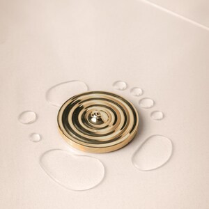 K&ouml;nnte beinhalten: Ein goldfarbener Metall-R&auml;ucherofen mit einem runden Design und einem kleinen Loch in der Mitte. Der R&auml;ucherofen ist von Wassertropfen auf einer wei&szlig;en Oberfl&auml;che umgeben.
