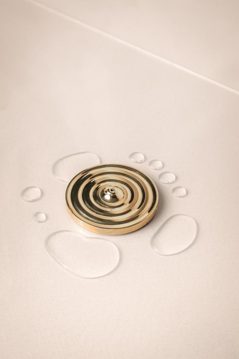 K&ouml;nnte beinhalten: Ein goldfarbener Metall-R&auml;ucherofen mit einem runden Design und einem kleinen Loch in der Mitte. Der R&auml;ucherofen ist von Wassertropfen auf einer wei&szlig;en Oberfl&auml;che umgeben.