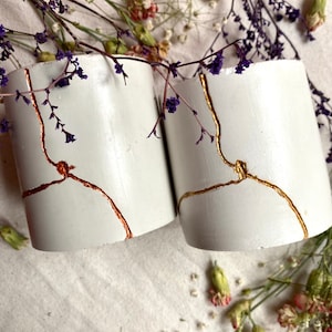 Peut inclure: Deux pots en béton blanc avec des motifs craquelés dorés et cuivrés. Les pots sont posés sur une surface en tissu blanc avec des fleurs séchées éparpillées autour.