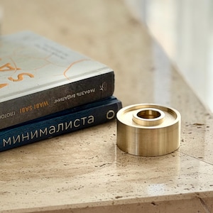 May include: A gold-colored candle holder with a circular design, featuring a central well. Two books are visible in the background. The candle holder is made of metal and sits on a light-colored surface.
