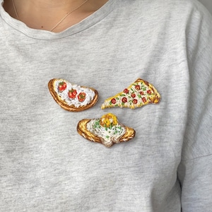May include: A gray t-shirt with three embroidered patches of different foods: a slice of pizza, a piece of toast with tomato and cheese, and a piece of toast with a fried egg.