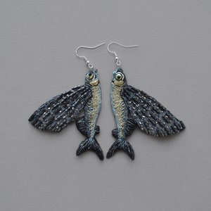 Puede incluir: Un par de pendientes con forma de pez. Cada pendiente presenta un pez con alas detalladas, en una combinación de colores azul y plateado con detalles en negro. Los pendientes cuelgan de ganchos plateados.