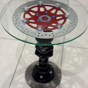 Puede incluir: Una mesa auxiliar única con una tapa de cristal transparente. La mesa presenta un rotor de freno de motocicleta rojo y plateado como base de la mesa, sostenido por una columna de metal negro y una base de rotor de freno. Un mueble creativo.