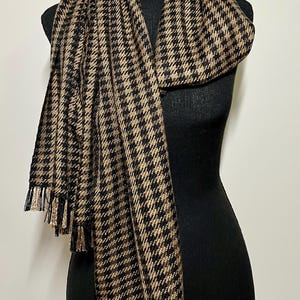 May include: A black and tan houndstooth scarf with fringe. The scarf is draped over a black mannequin. The pattern is a classic houndstooth design, with alternating black and tan squares. The fringe is a mix of black and tan threads.
