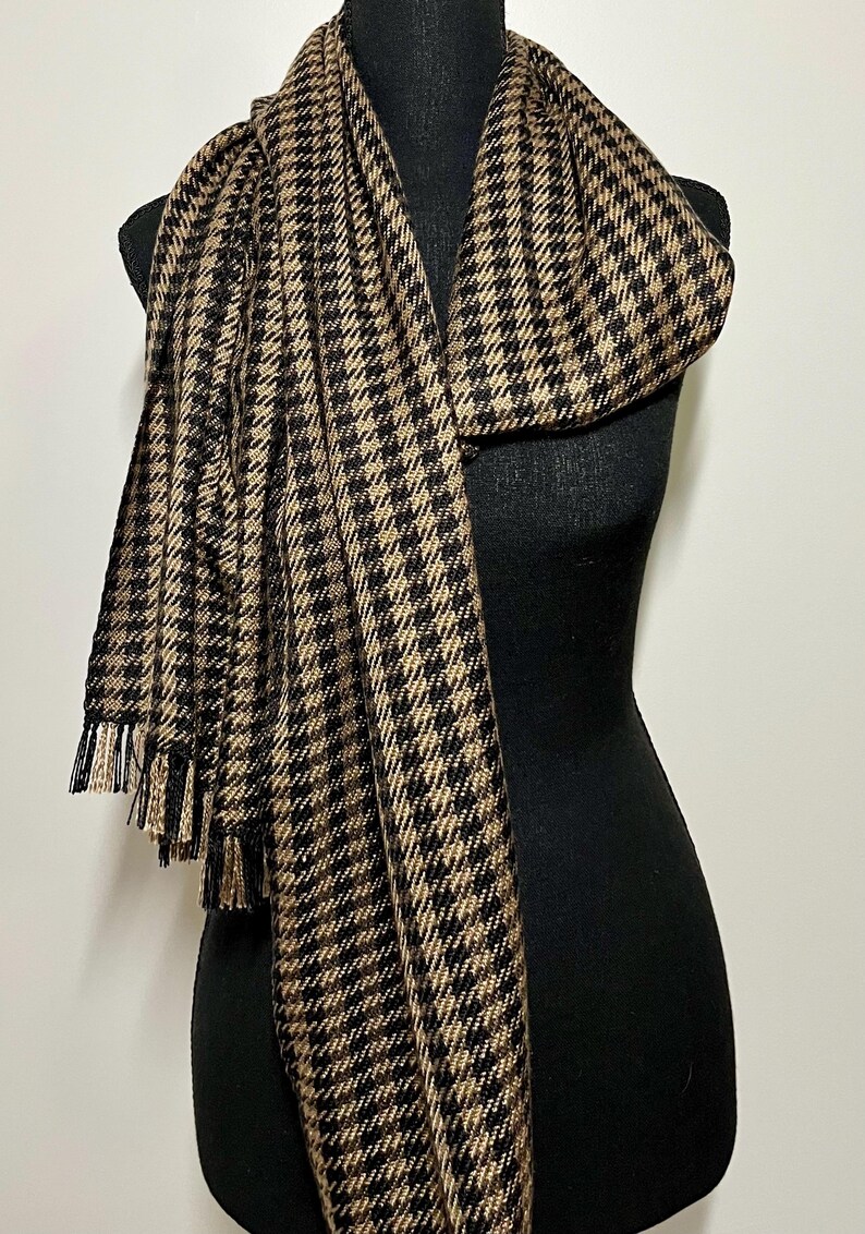 May include: A black and tan houndstooth scarf with fringe. The scarf is draped over a black mannequin. The pattern is a classic houndstooth design, with alternating black and tan squares. The fringe is a mix of black and tan threads.