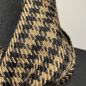 May include: Close-up of a houndstooth scarf in black and tan. The fabric is tightly woven, showcasing a classic pattern. The scarf is draped over a black form, highlighting its texture and design. A stylish accessory.