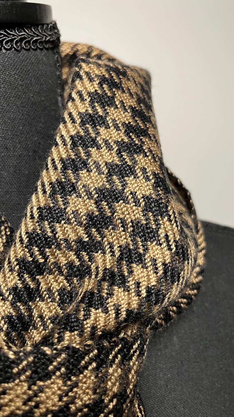May include: Close-up of a houndstooth scarf in black and tan. The fabric is tightly woven, showcasing a classic pattern. The scarf is draped over a black form, highlighting its texture and design. A stylish accessory.