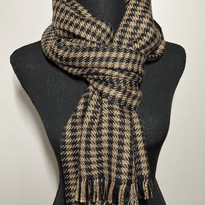 May include: A houndstooth patterned scarf in black and tan, draped over a black mannequin. The scarf has a fringed edge and is made of a woven fabric. The scarf is a neutral accessory.