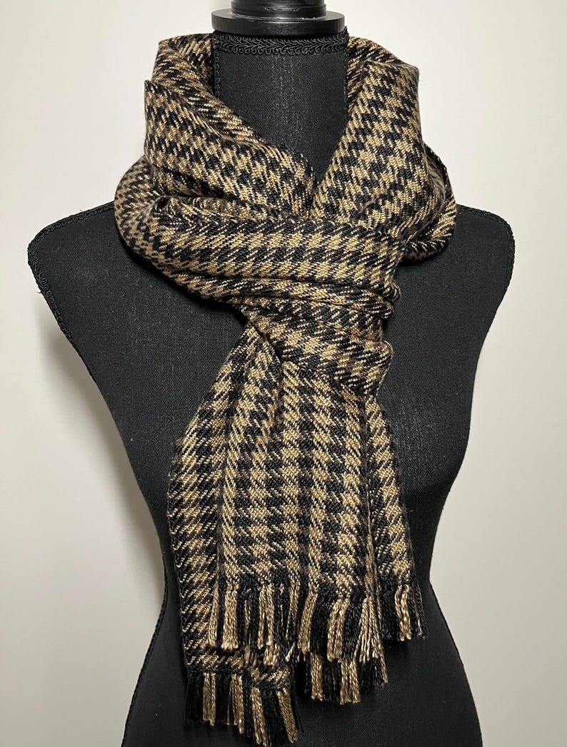 May include: A houndstooth patterned scarf in black and tan, draped over a black mannequin. The scarf has a fringed edge and is made of a woven fabric. The scarf is a neutral accessory.