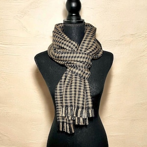 May include: A black and tan houndstooth scarf with fringe. The scarf is draped over a black mannequin. The scarf has a classic pattern and is made of a woven material. The scarf is a stylish accessory.