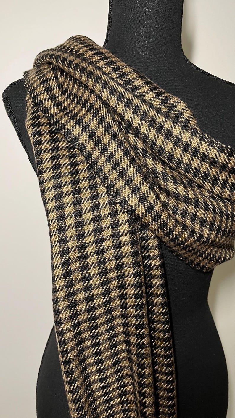 May include: A black and tan houndstooth pattern scarf draped over a black mannequin. The scarf is made of a soft, woven material. The pattern is a classic design, with small, repeating squares.