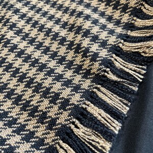 May include: Close-up of a black and tan houndstooth pattern textile with fringed edges. The fabric has a classic, woven design. The fringe is tan and black.