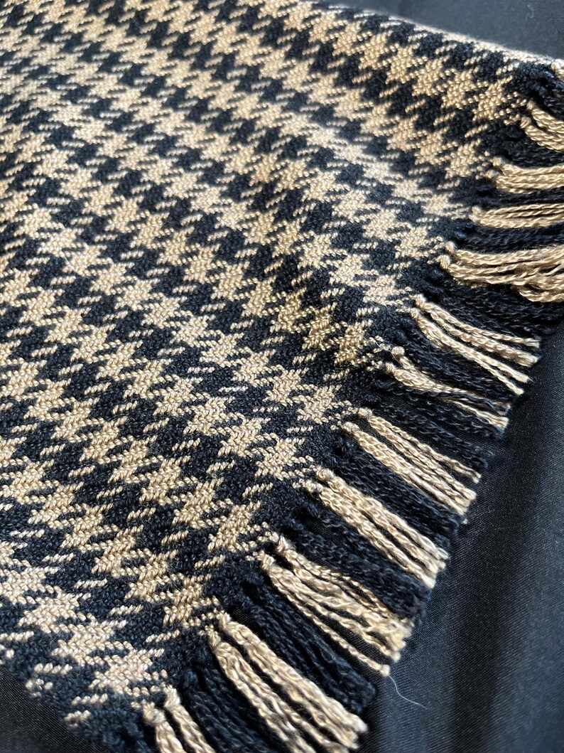 May include: Close-up of a black and tan houndstooth pattern textile with fringed edges. The fabric has a classic, woven design. The fringe is tan and black.